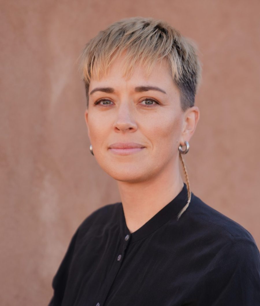 Close-up portrait of a person with short, textured blonde hair and darker roots, wearing small hoop earrings and a black collared shirt, looking calmly at the camera against a warm-toned background.