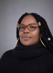 Portrait of a woman with long braided hair, wearing red cat-eye glasses with gold accents, large hoop earrings, and a black turtleneck, facing the camera against a plain gray background.