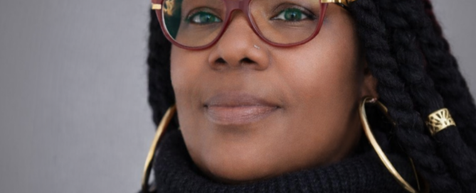 Portrait of a woman with long braided hair, wearing red cat-eye glasses with gold accents, large hoop earrings, and a black turtleneck, facing the camera against a plain gray background.