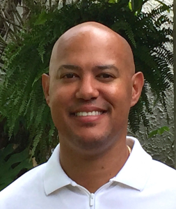  Portrait of a bald man smiling at the camera, wearing a white collared shirt, standing outdoors in front of green foliage.