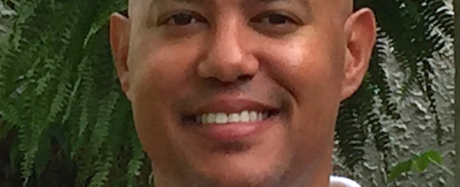 Portrait of a bald man smiling at the camera, wearing a white collared shirt, standing outdoors in front of green foliage.