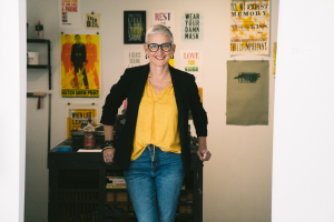  Full-body portrait of a person with short gray hair and glasses, wearing a yellow top, black blazer, and jeans, standing with hands resting on a table in a studio with colorful posters on the wall.