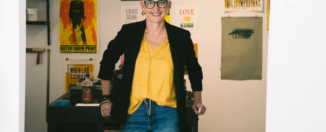 Full-body portrait of a person with short gray hair and glasses, wearing a yellow top, black blazer, and jeans, standing with hands resting on a table in a studio with colorful posters on the wall.