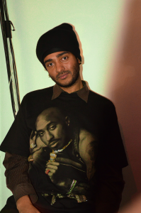 Portrait of a person wearing a black beanie and a black graphic T-shirt featuring an image of Tupac, looking at the camera under warm indoor lighting.