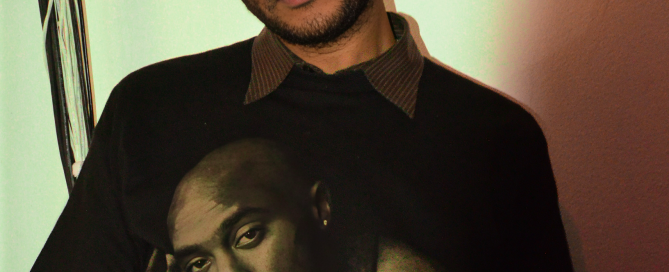 Portrait of a person wearing a black beanie and a black graphic T-shirt featuring an image of Tupac, looking at the camera under warm indoor lighting.