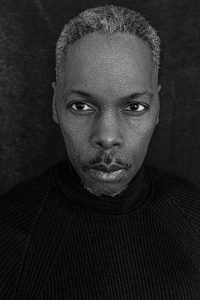 Black-and-white close-up portrait of a person with short textured hair and a mustache, wearing a dark sweater, looking directly at the camera against a dark backdrop.