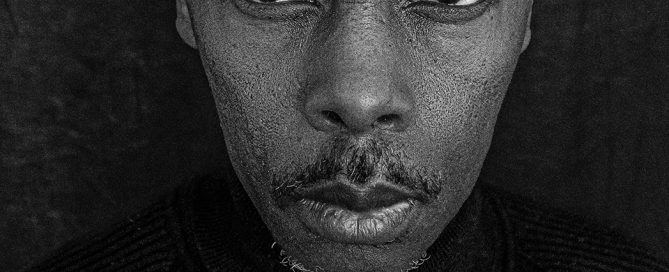 Black-and-white close-up portrait of a person with short textured hair and a mustache, wearing a dark sweater, looking directly at the camera against a dark backdrop.