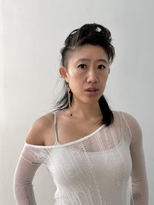 Portrait of a person with partially shaved sides and longer dark hair swept to one side, wearing a sheer white off-the-shoulder top and long dangling earrings, looking directly at the camera against a plain white background.