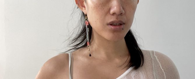 Portrait of a person with partially shaved sides and longer dark hair swept to one side, wearing a sheer white off-the-shoulder top and long dangling earrings, looking directly at the camera against a plain white background.